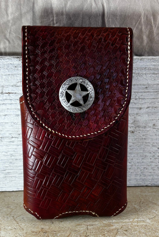 A dark reddish-brown leather phone pouch with a woven basket-style embossed pattern and a silver-toned star-shaped snap button, standing upright against a rustic white wood panel and beige stone surface.